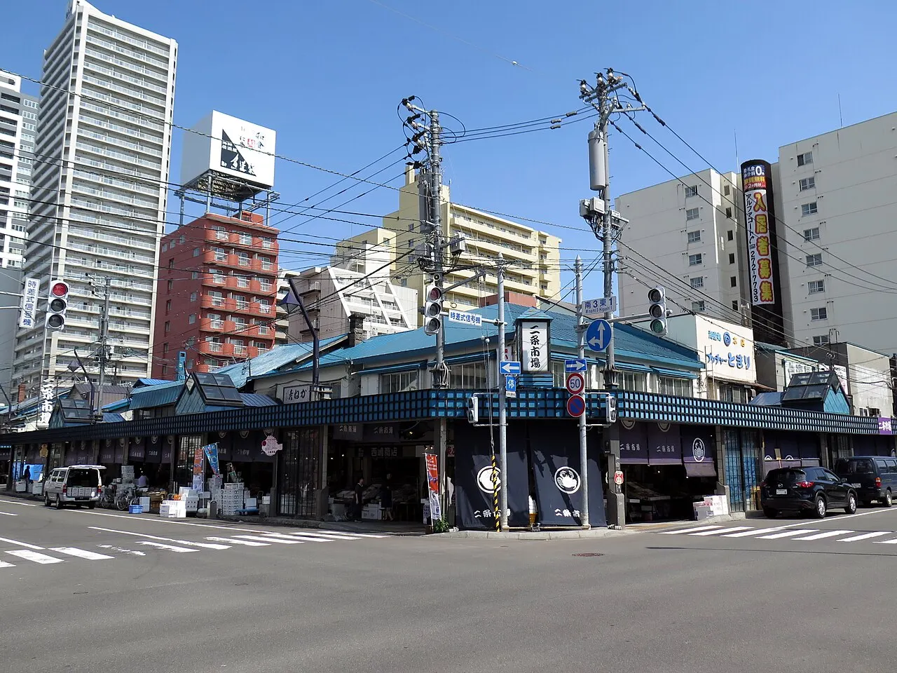A row of single-storey shops with blue roofs and dark awnings fills a street corner, with boxes on the pavement, utility poles and tall buildings behind.