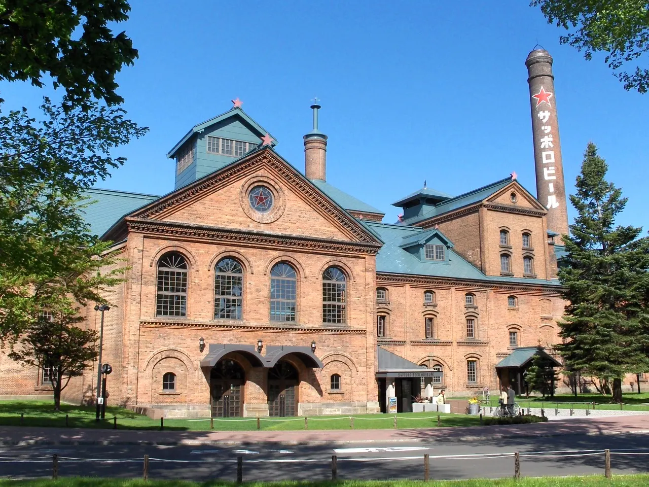 A brick building with a triangular pediment, arched windows and green roofs, beside a tall chimney with a red star and Japanese lettering, under a blue sky.
