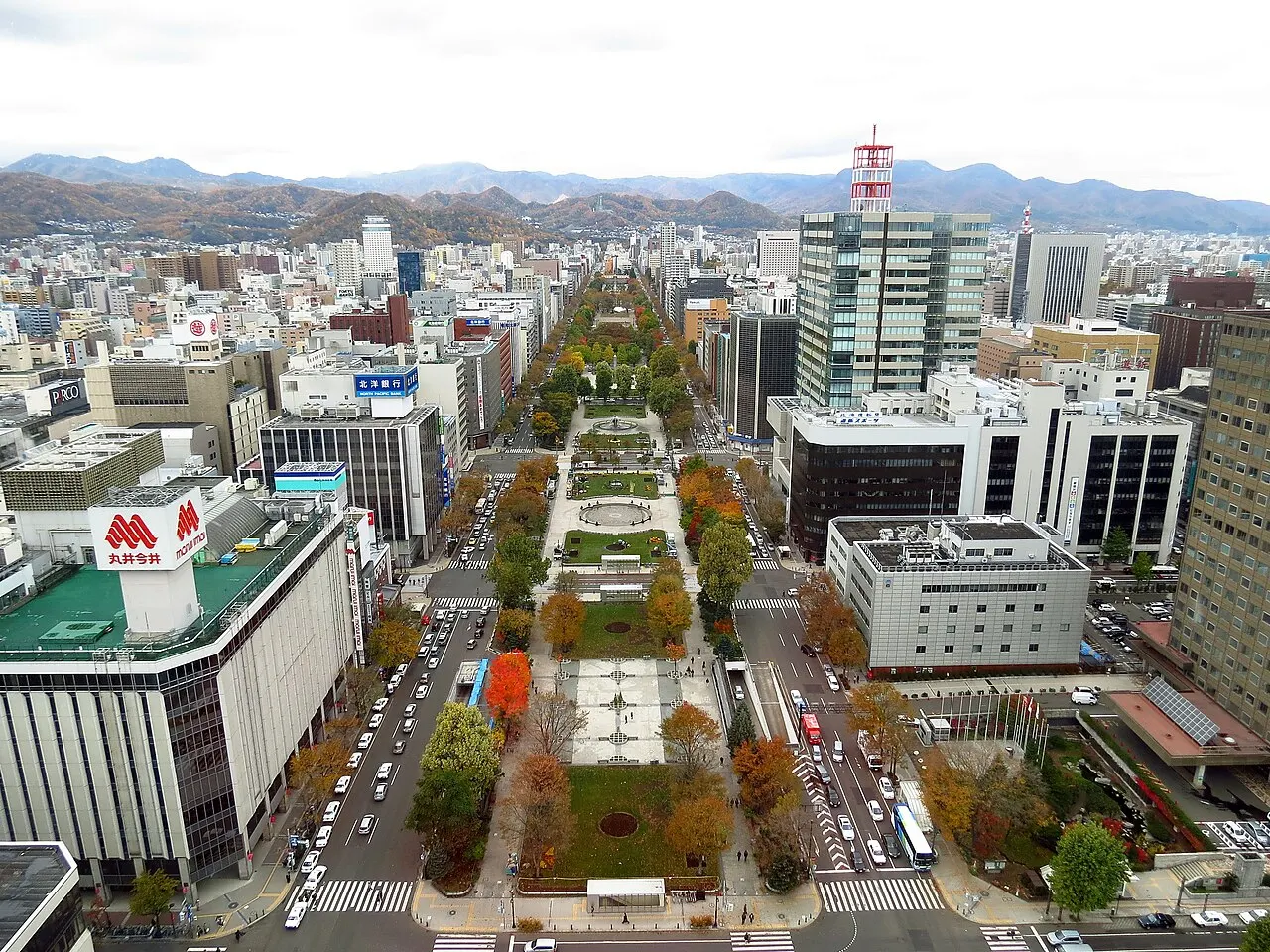 High view of a strip of gardens with autumn trees, lawns and paths cutting through the city between two wide avenues, with buildings on both sides and mountains in the background.
