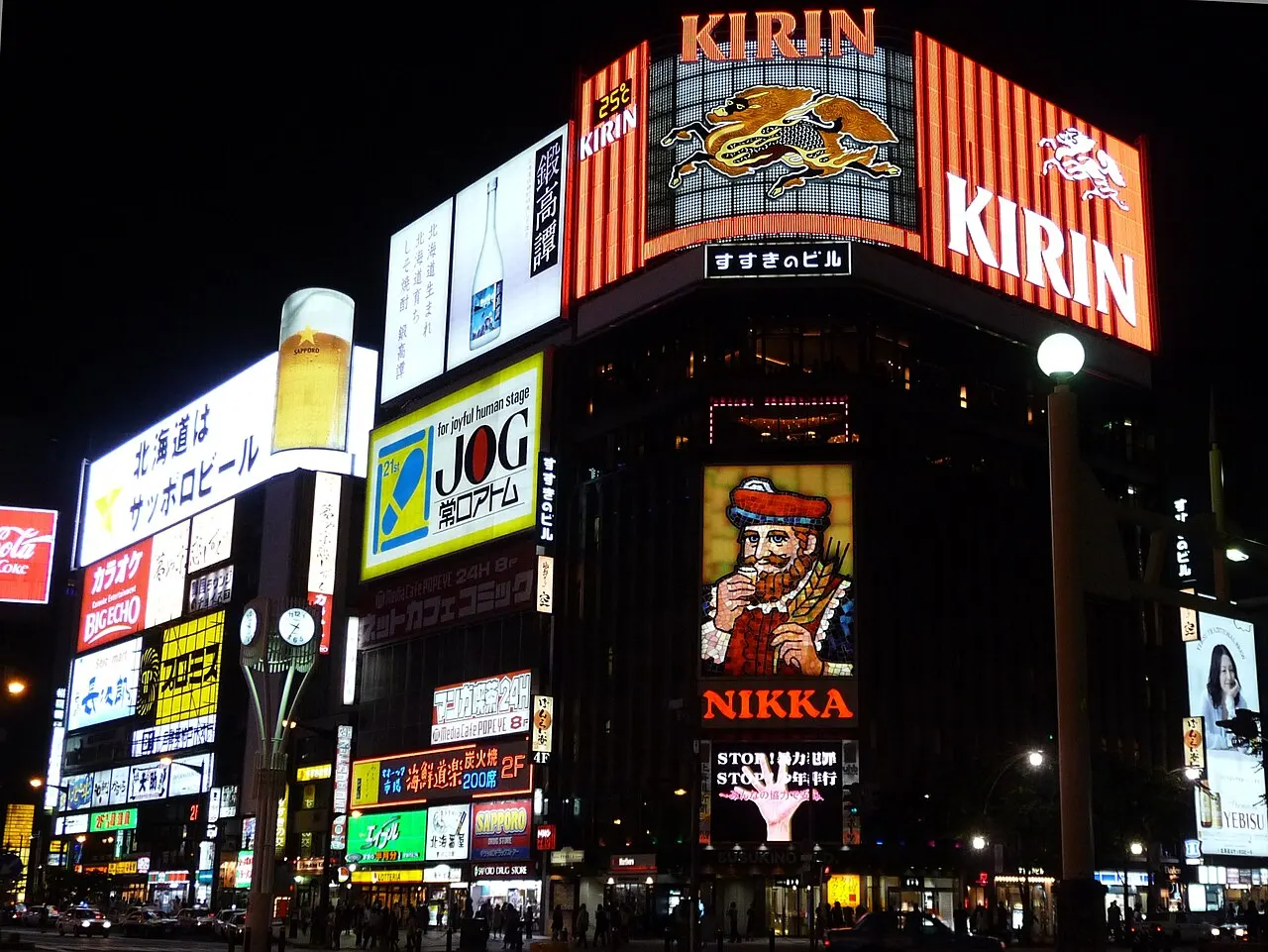 At night, corner buildings covered in colourful illuminated signs, among them a large one showing a bearded man, above a street with pedestrians.