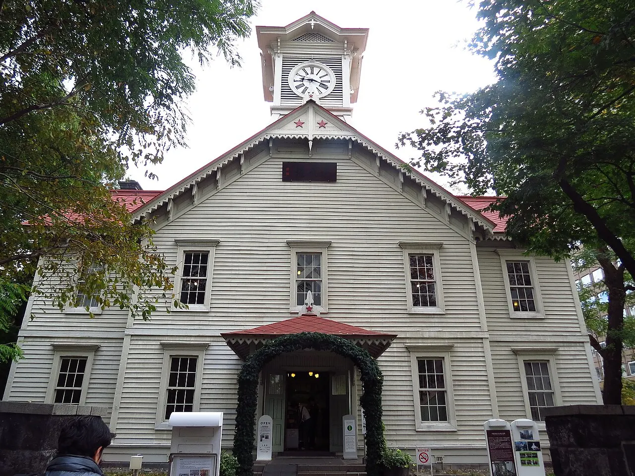 Edificio de madera pintado de blanco, con techo rojo, ventanas de guillotina y una torre con reloj en lo alto, enmarcado por árboles.