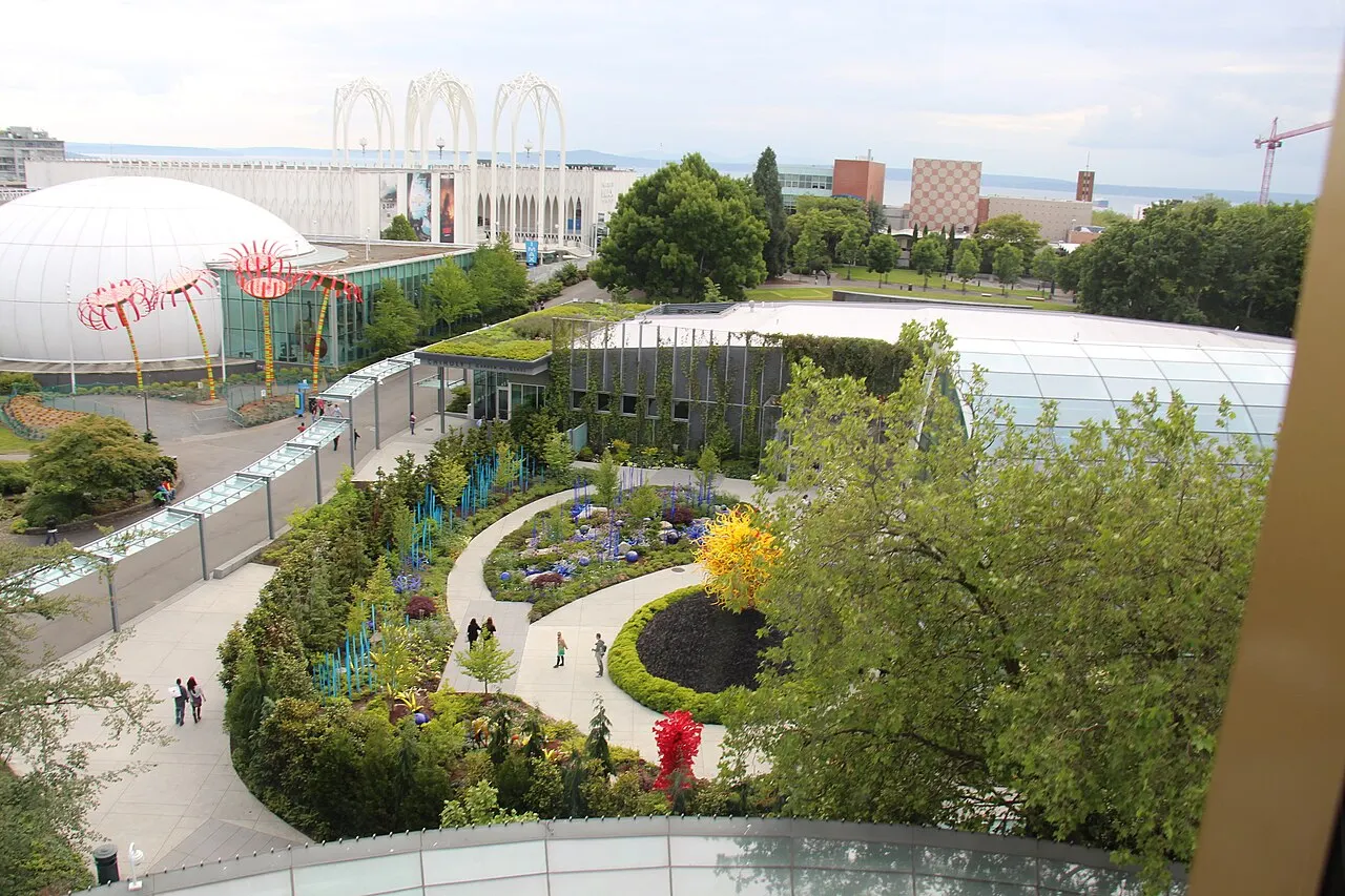 Vista de cima do jardim do Chihuly Garden and Glass, com canteiros sinuosos, árvores e as estufas ao fundo.