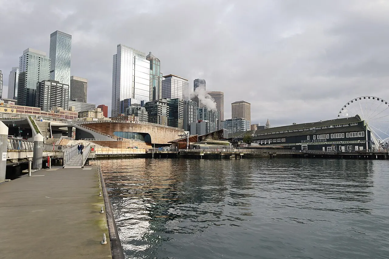 Orla de Seattle com o prédio do Seattle Aquarium à beira da água, arranha-céus e uma roda-gigante ao fundo, sob céu nublado.