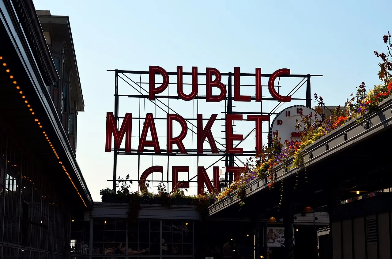 Enseigne rouge au néon du Pike Place Market avec une horloge ronde à côté, des paniers de fleurs colorées sur l’auvent de l’entrée, au crépuscule.