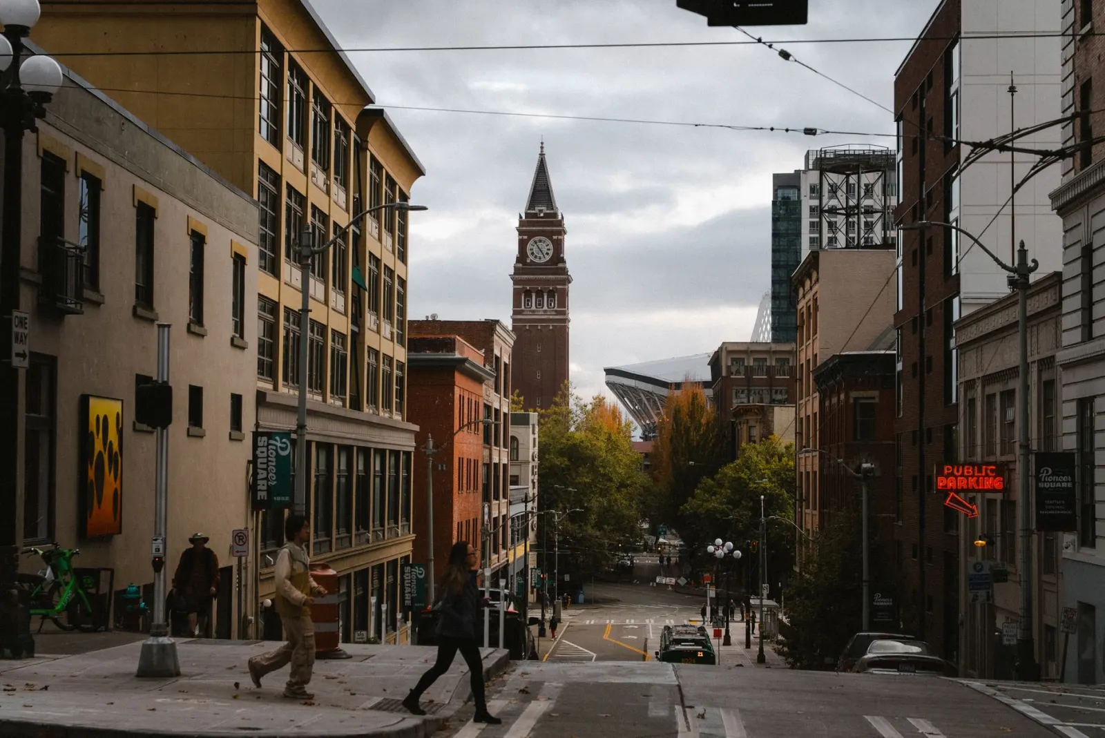 Rua de Pioneer Square, em Seattle, descendo entre prédios de tijolo e alvenaria antigos com galhardetes do bairro, fios de trólebus cruzando o alto e a torre de relógio de tijolo da King Street Station ao fundo, sob céu cinzento.
