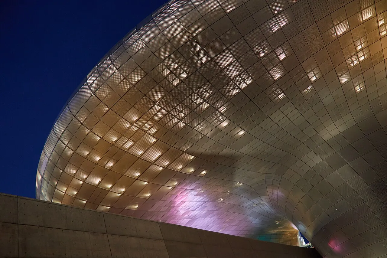 Superfície curva de painéis metálicos do Dongdaemun Design Plaza, iluminada ao entardecer, vista de baixo.