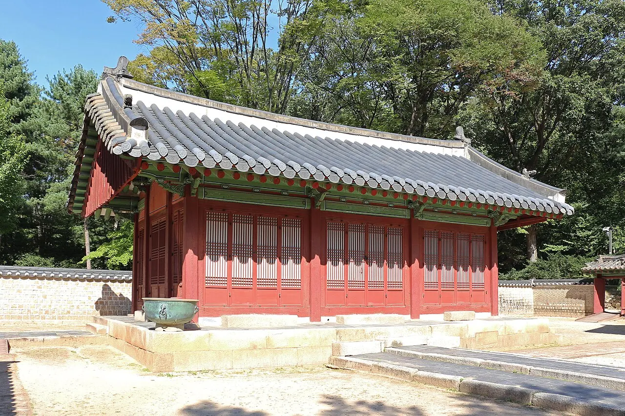 Pavilhão comprido de colunas vermelhas e telhado cinza, num pátio de pedra entre árvores.