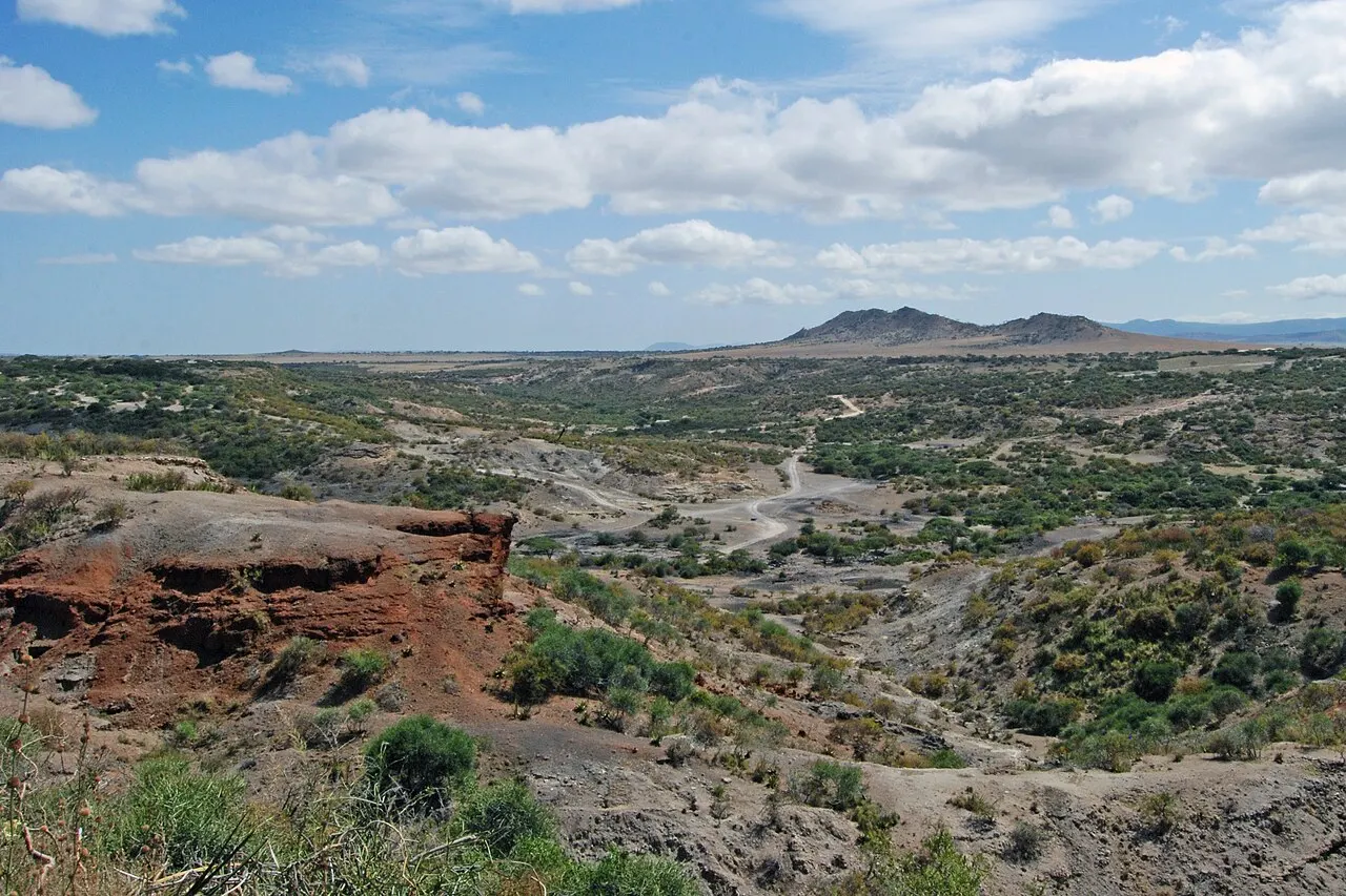 Uma garganta seca e erodida, com um barranco de camadas vermelhas à esquerda, encostas cinzentas com arbustos verdes, uma estrada de terra no fundo e um morro escuro no horizonte sob céu com nuvens.