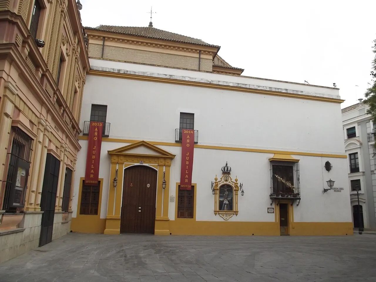 Fachada caiada de branco e ocre do Museu de Belas Artes de Sevilha, antigo convento, com portal de madeira sob frontão triangular, dois estandartes vermelhos com a palavra “museo”, um retábulo de azulejo na parede e a praça vazia em primeiro plano.