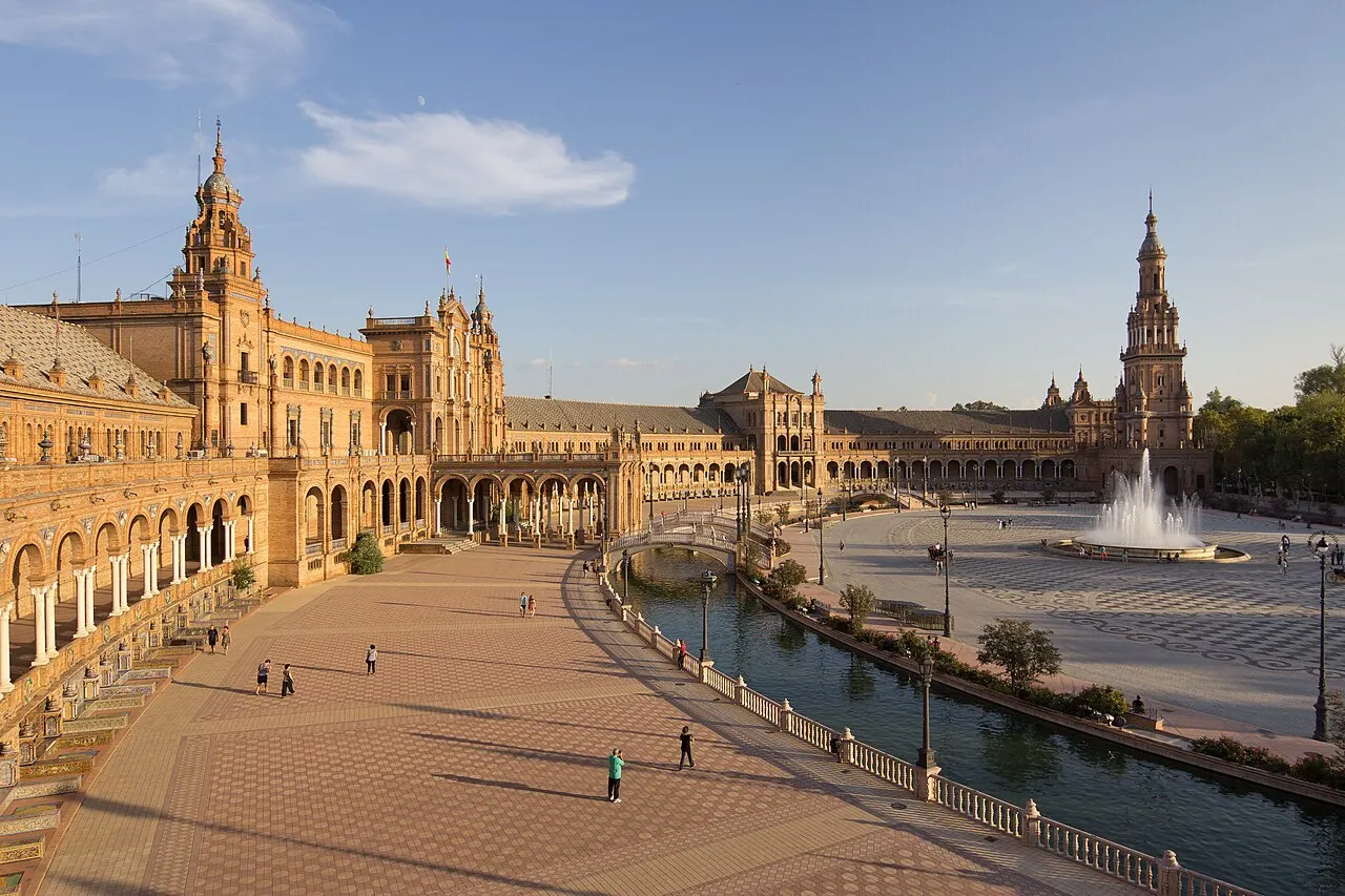 A Plaza de España, com o edifício semicircular de tijolo e torres, o canal e pessoas caminhando na praça.