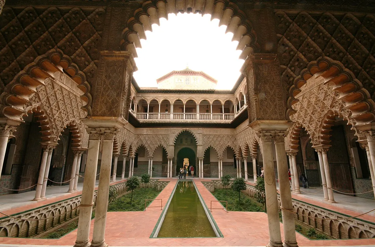 Pátio interno do Real Alcázar de Sevilha, com arcos mouriscos ornamentados, colunas de mármore e um espelho d'água estreito ao centro.