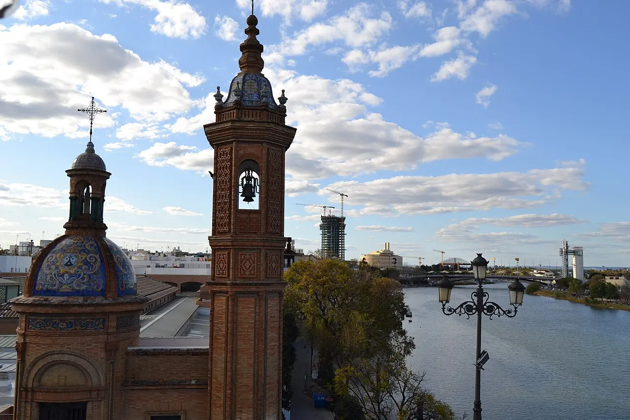 Duas torres sineiras com cúpulas de azulejo junto ao rio Guadalquivir, guindastes de obra e prédios ao fundo, sob céu com nuvens esparsas.