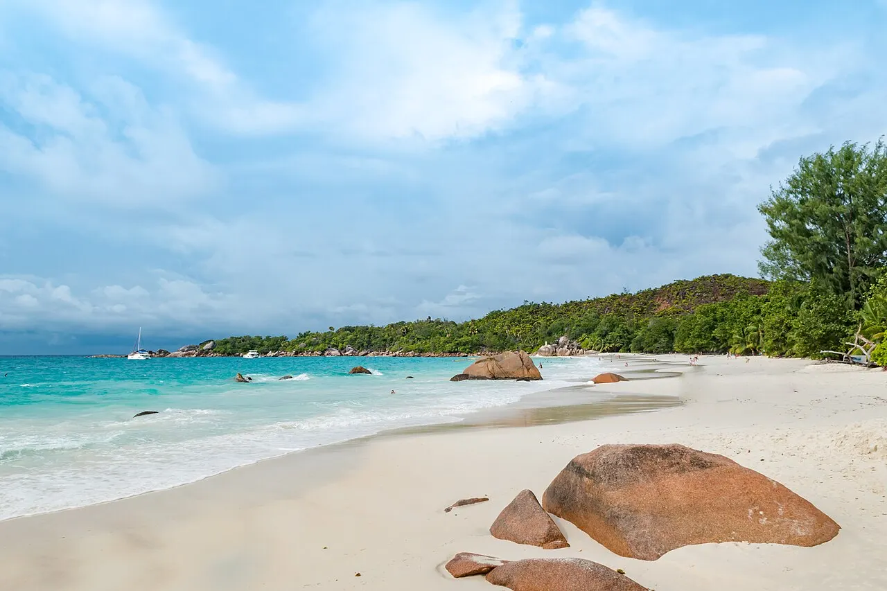 Plage de sable blanc avec des rochers orangés au bord de l'eau turquoise et une colline verte au fond.