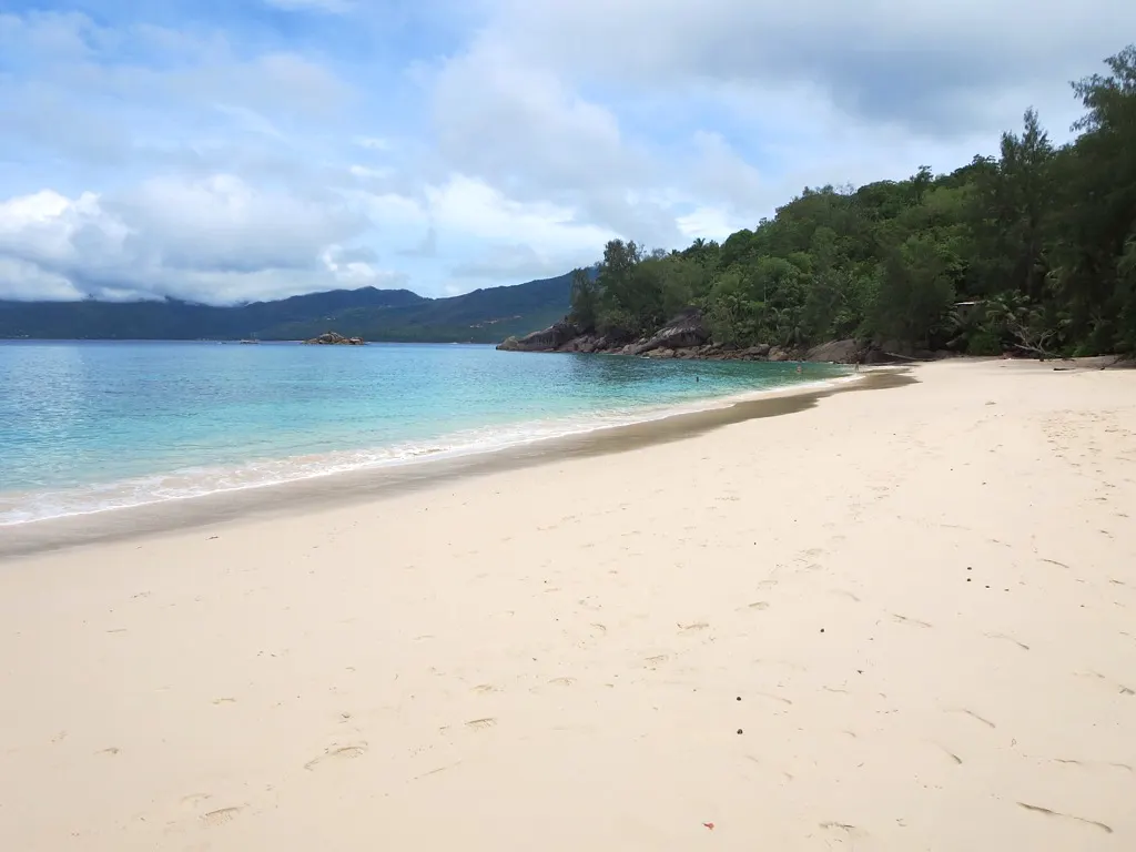 Bande de sable clair en arc au bord d'une eau bleue et calme, avec des arbres sur la pointe de la crique et des montagnes au loin.