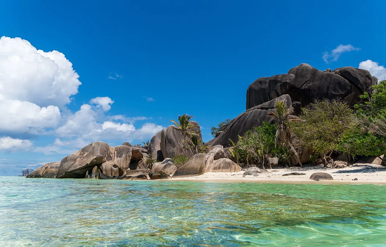 De gros rochers de granit arrondis sur une bande de sable blanc, avec de la végétation verte derrière et une eau peu profonde et transparente au premier plan.