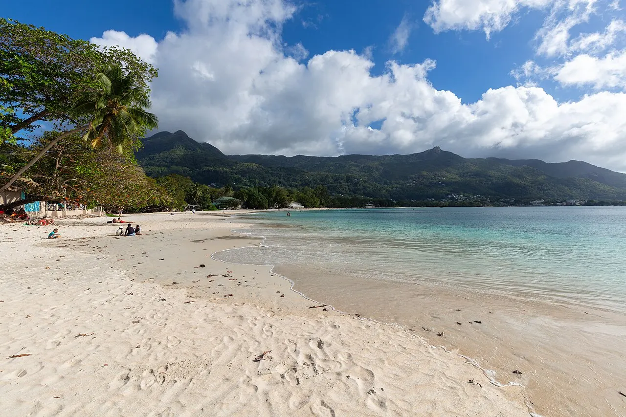 Longue plage de sable clair à l'eau calme et peu profonde, des arbres à gauche, quelques personnes sur le sable et des montagnes vertes au fond de la baie.