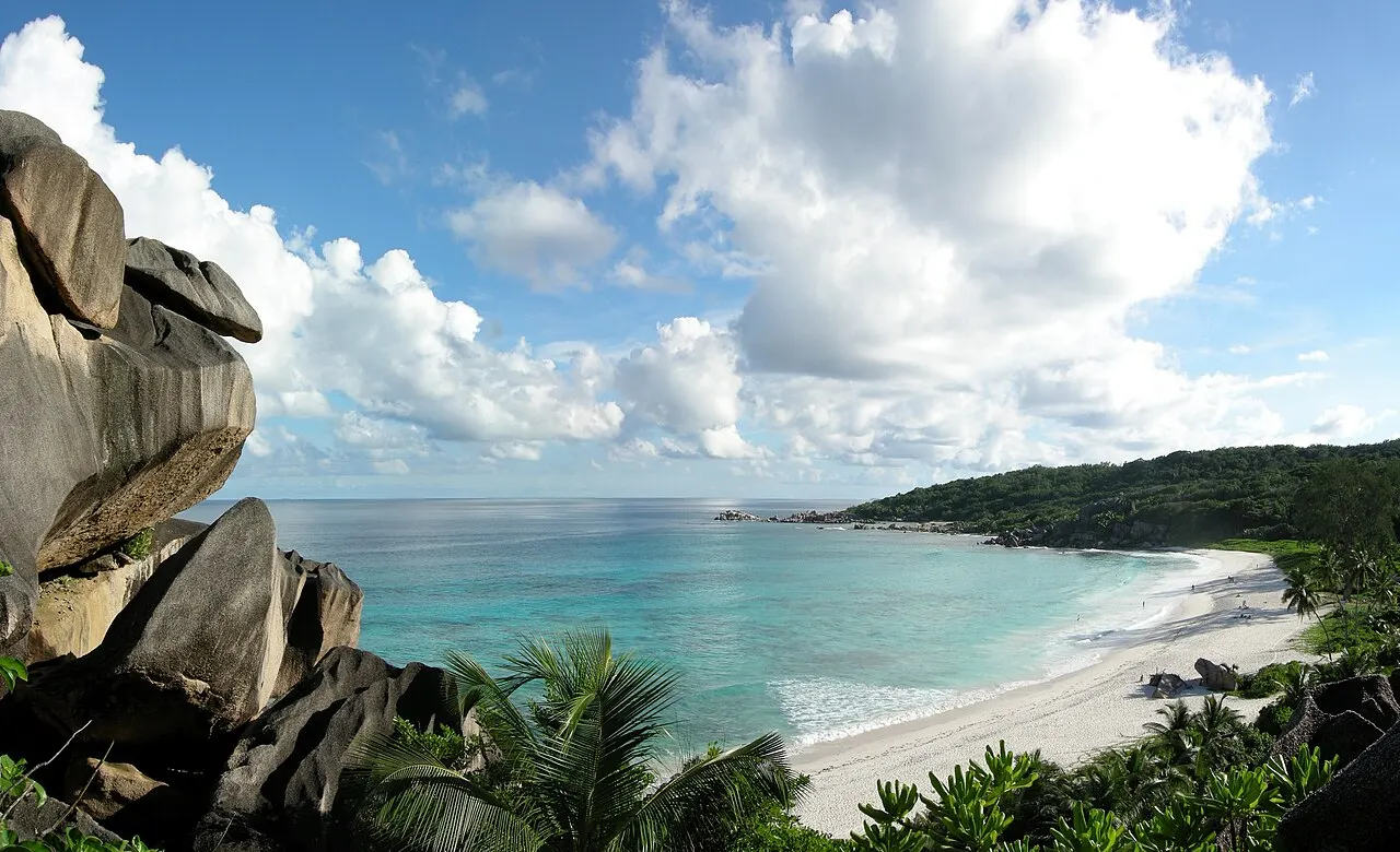 Large baie de sable blanc et de mer bleue avec des vagues, vue d'en haut entre des rochers de granit à gauche et la végétation autour.