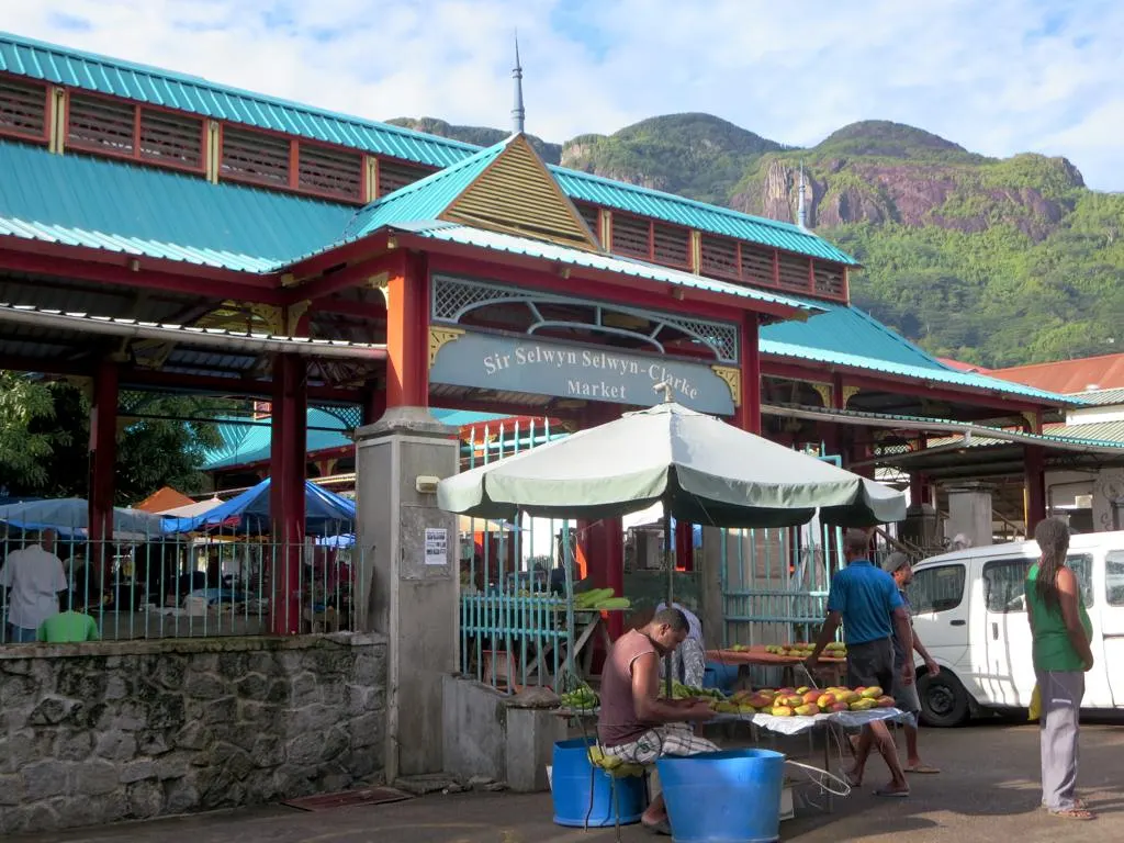 La façade du marché au toit vert et aux piliers rouges, l'enseigne au-dessus de l'entrée, un vendeur de fruits sous un parasol et la colline verte derrière.