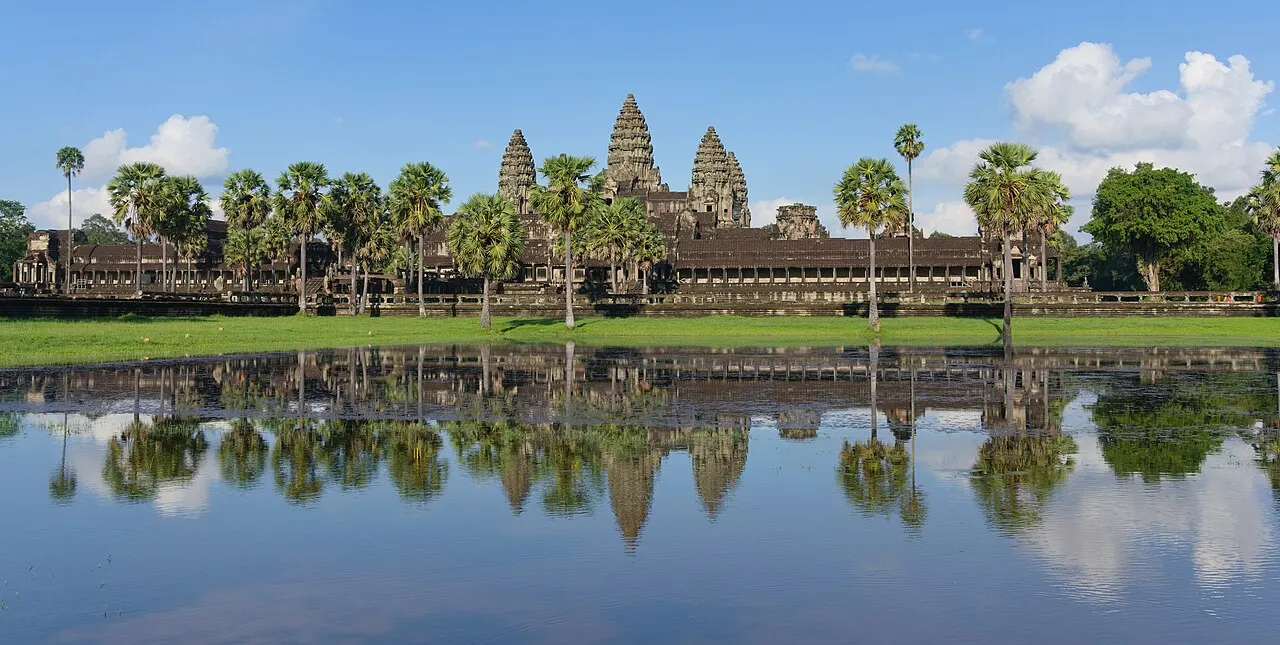 As torres em forma de flor de lótus de Angkor Wat entre palmeiras, refletidas num espelho d'água calmo, sob céu azul com nuvens.