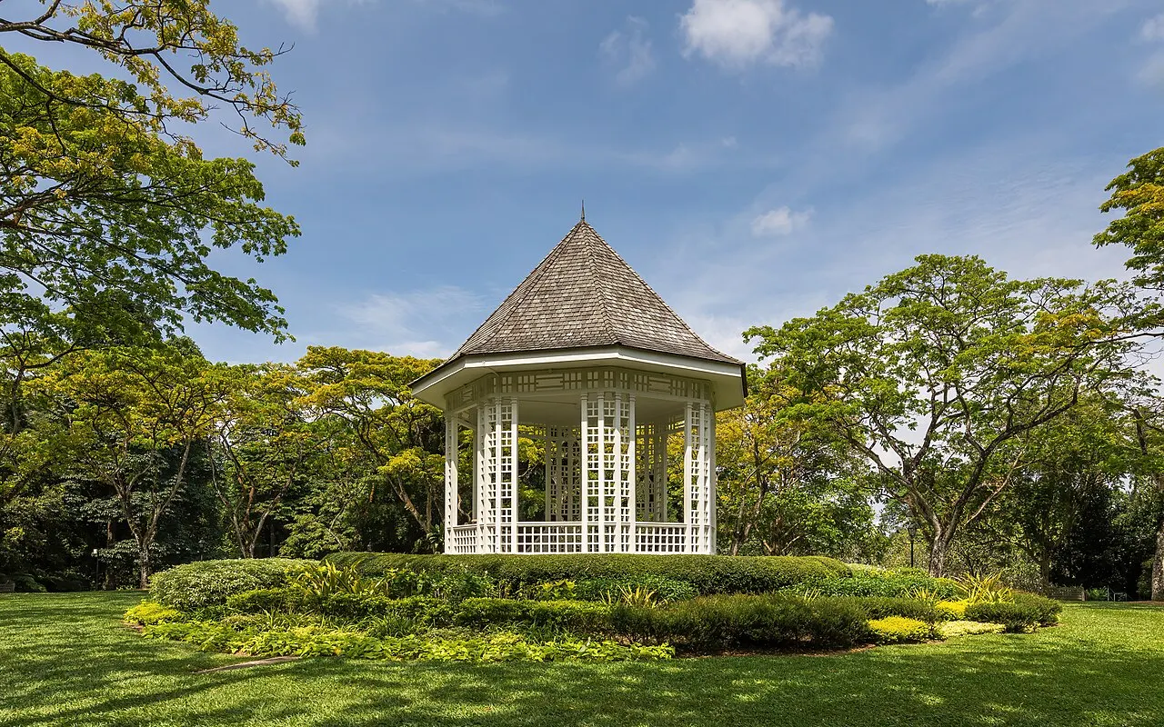 Coreto branco octogonal de telhado alto, num gramado cercado de árvores.