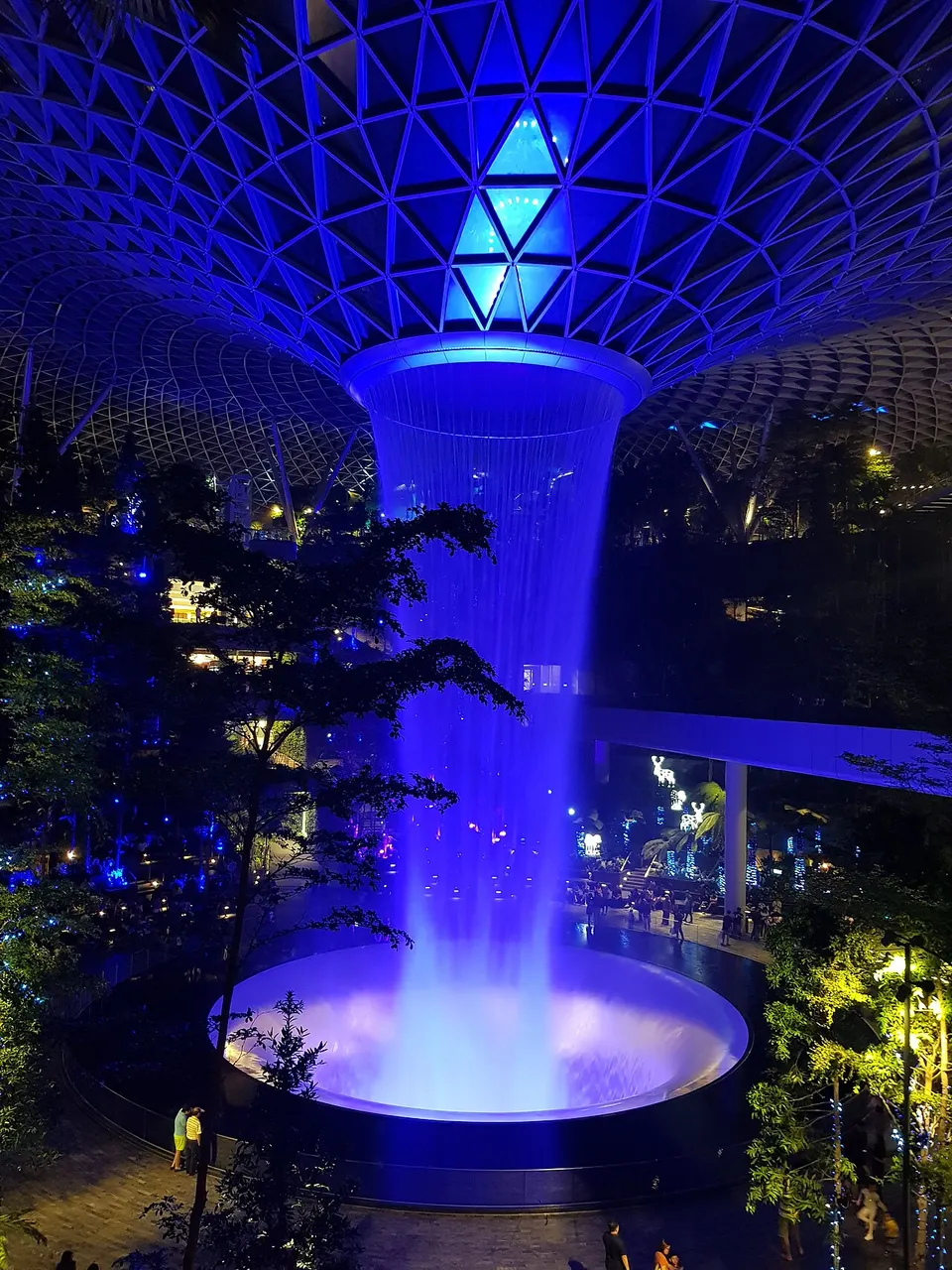 Cone de água iluminado de azul caindo dentro de um átrio de vidro, à noite.