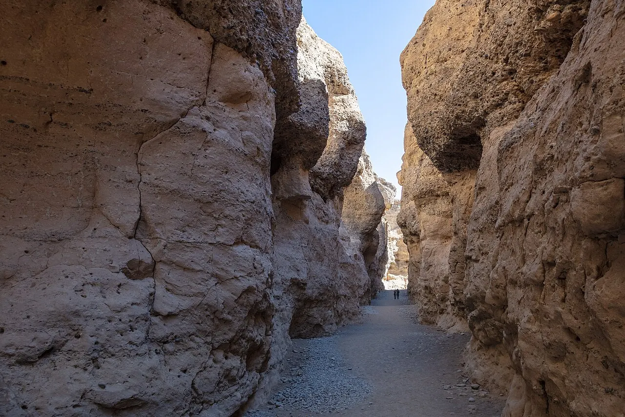The dry gravel floor of Sesriem Canyon between high walls of pale, eroded sedimentary rock, with a strip of blue sky above and two small figures in the distance.