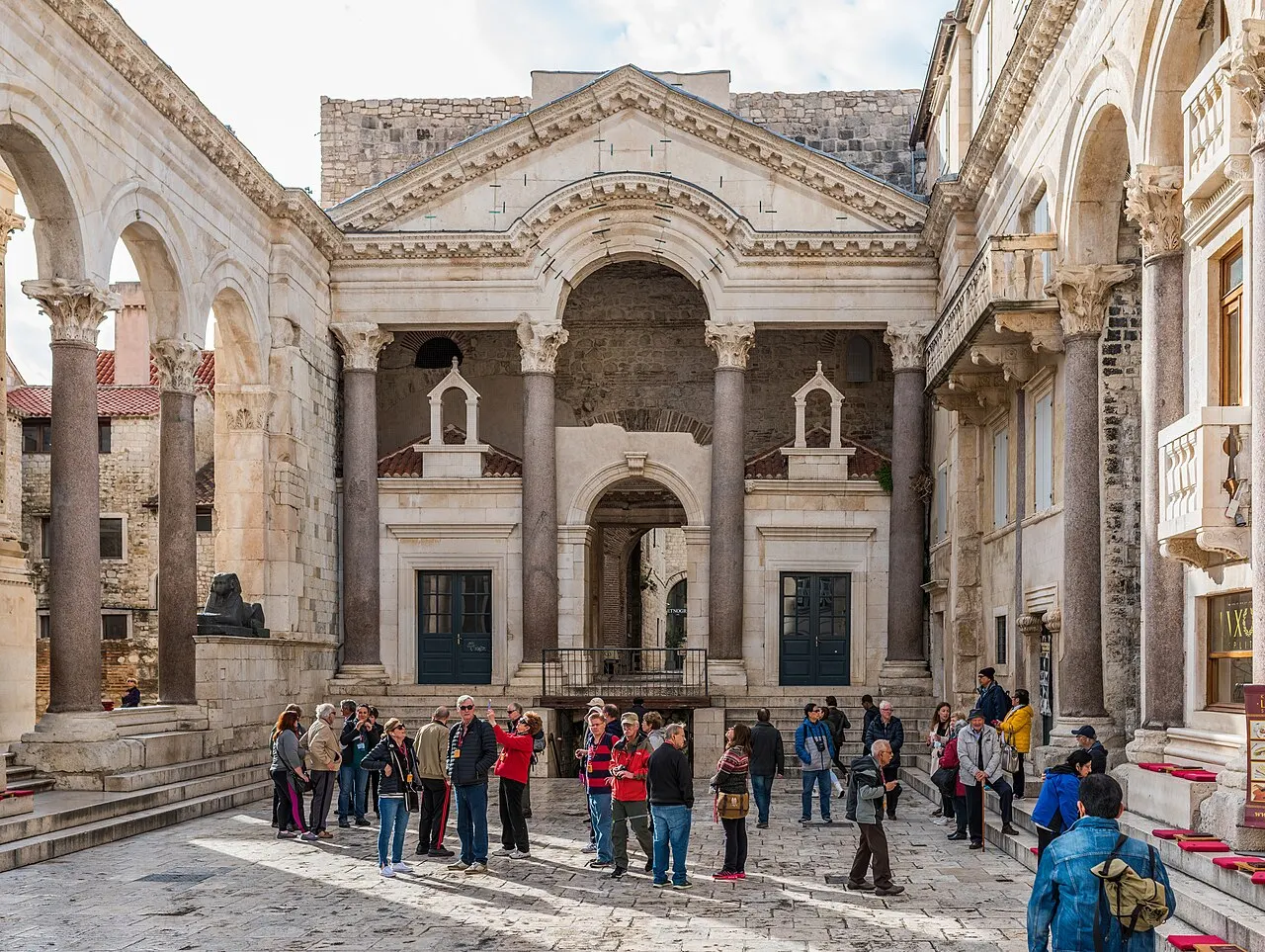 O Peristilo do Palácio de Diocleciano, com colunas e arcos dos dois lados e o pórtico de frontão ao fundo, cheio de visitantes.