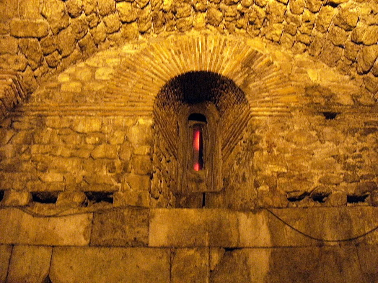 Uma parede de pedra dos porões do palácio sob luz amarela, com um arco de tijolos em leque em volta de uma pequena janela.
