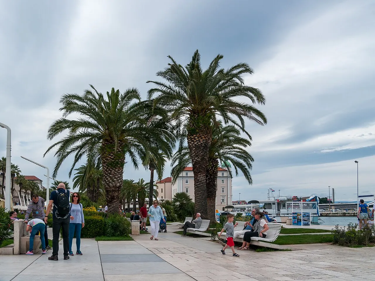 O calçadão de pedra clara da Riva com duas palmeiras altas, bancos brancos e pessoas passeando, num dia nublado.