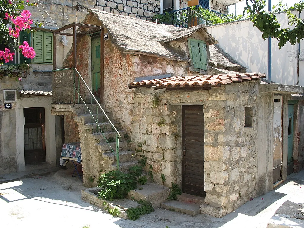 Uma casa antiga de pedra do Veli Varoš com escada externa de ferro, telhado de lajes, porta de madeira e flores na janela.