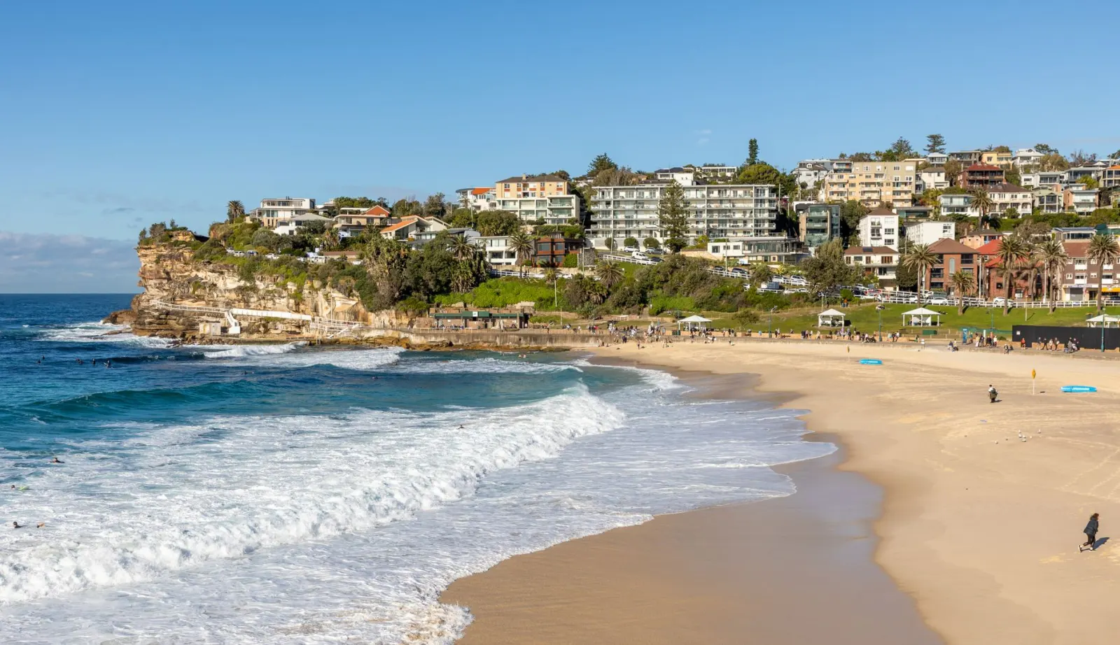 Praia em meia-lua no caminho costeiro do leste de Sydney: ondas brancas quebrando na areia clara, uma ponta de arenito com piscina de mar à esquerda e prédios baixos e casas subindo a encosta à direita.