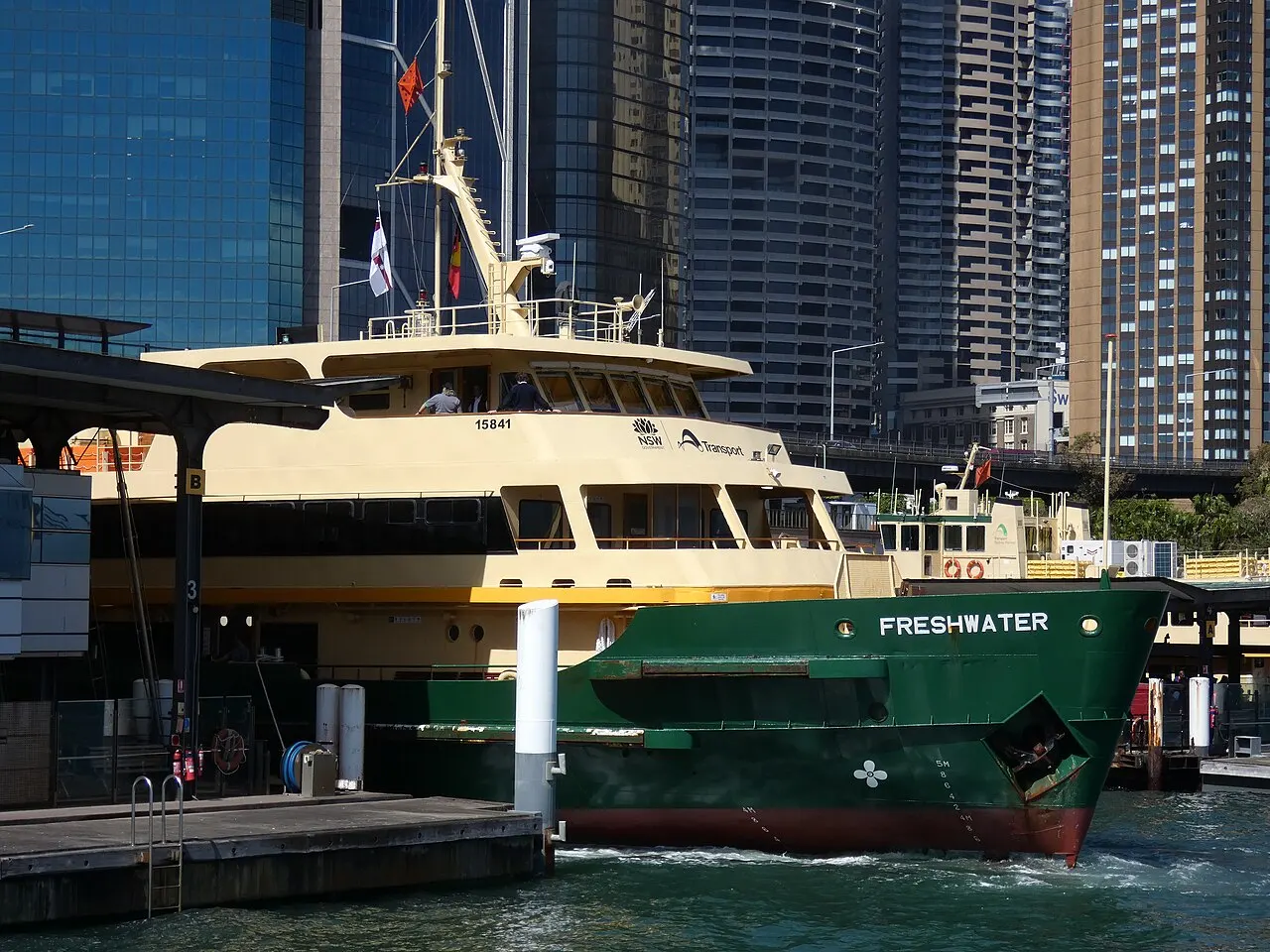 Balsa verde e creme chamada Freshwater atracada em Circular Quay, com arranha-céus de vidro ao fundo sob luz de sol forte.