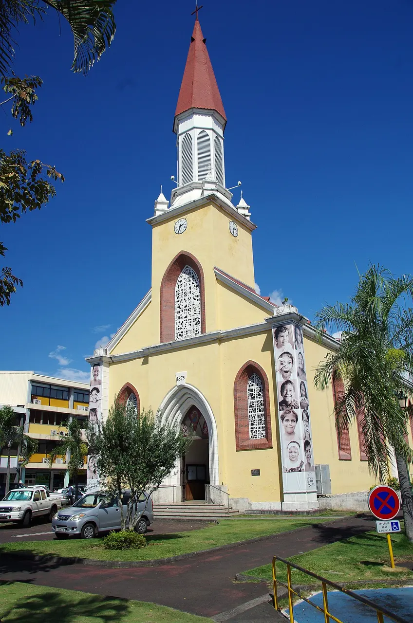 Catedral Notre-Dame de Papeete, de paredes amarelas e torre com agulha vermelha, sob céu azul.
