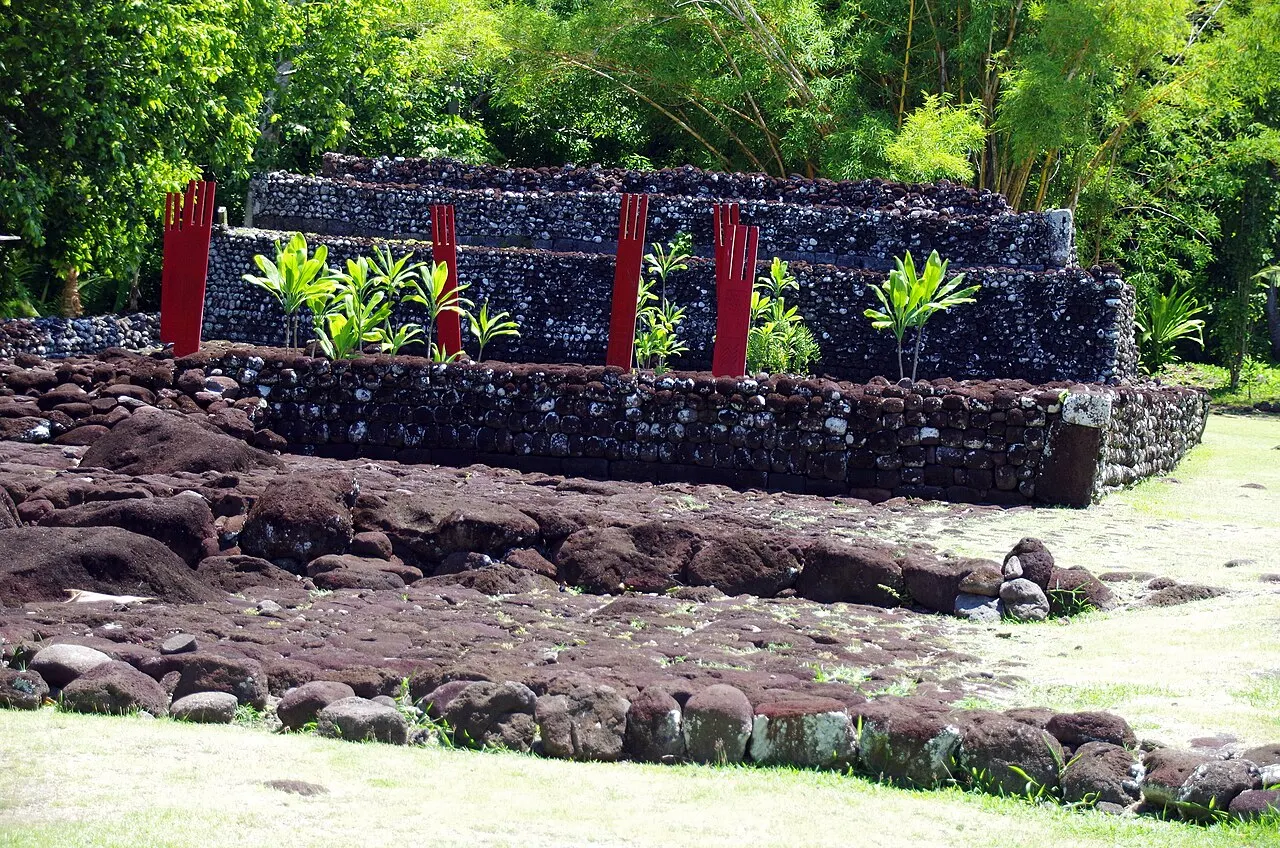 Plataforma escalonada de pedras vulcânicas do Marae Arahurahu, com postes vermelhos cerimoniais e plantas verdes ao longo do muro, cercada de vegetação densa.