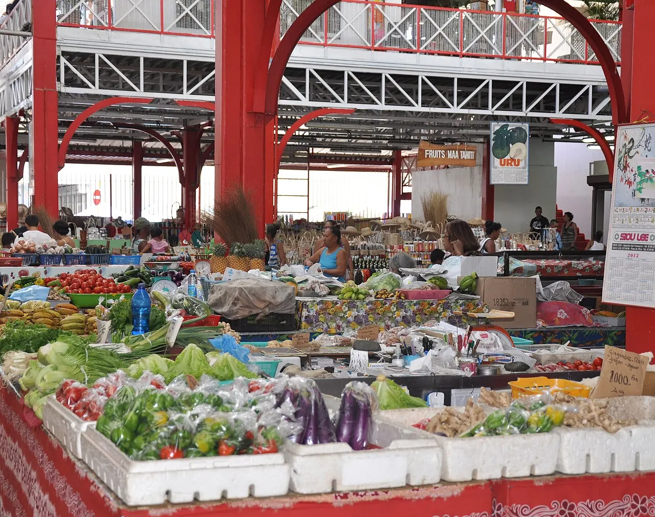 Interior do Marché de Papeete, com estrutura de ferro vermelha, barracas cheias de frutas e verduras e pessoas circulando entre os corredores.