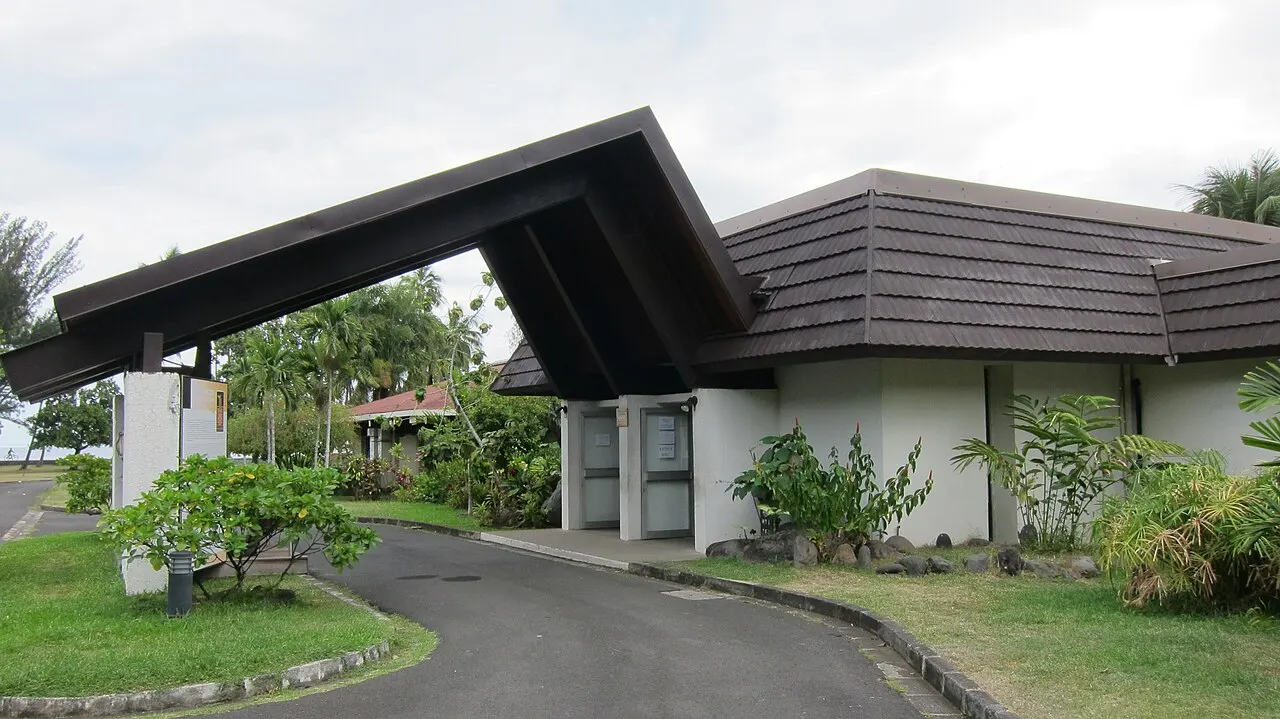 Entrada de um prédio baixo com telhado de duas águas muito inclinado em estilo polinésio, cercado de jardim tropical, sob céu nublado.