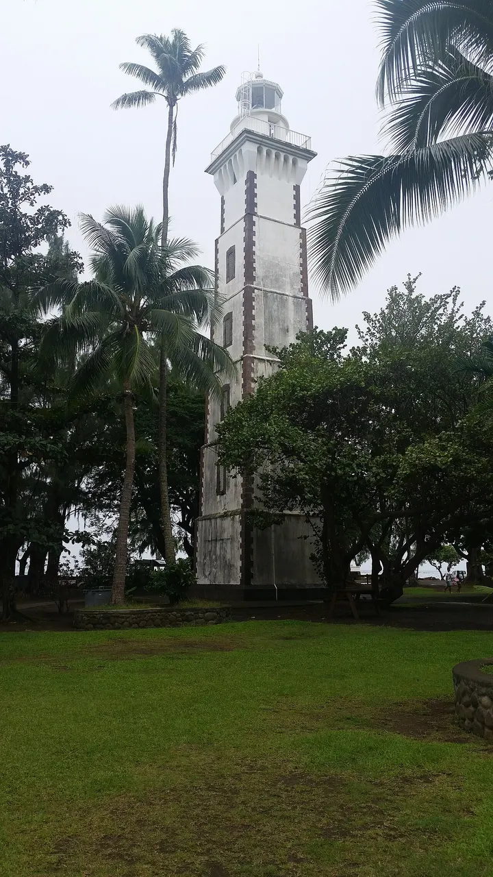 Torre branca de pedra do farol da Pointe Vénus, com cantos de tijolo aparente e lanterna no topo, entre coqueiros altos e árvores, sobre um gramado, com o mar aparecendo à esquerda.