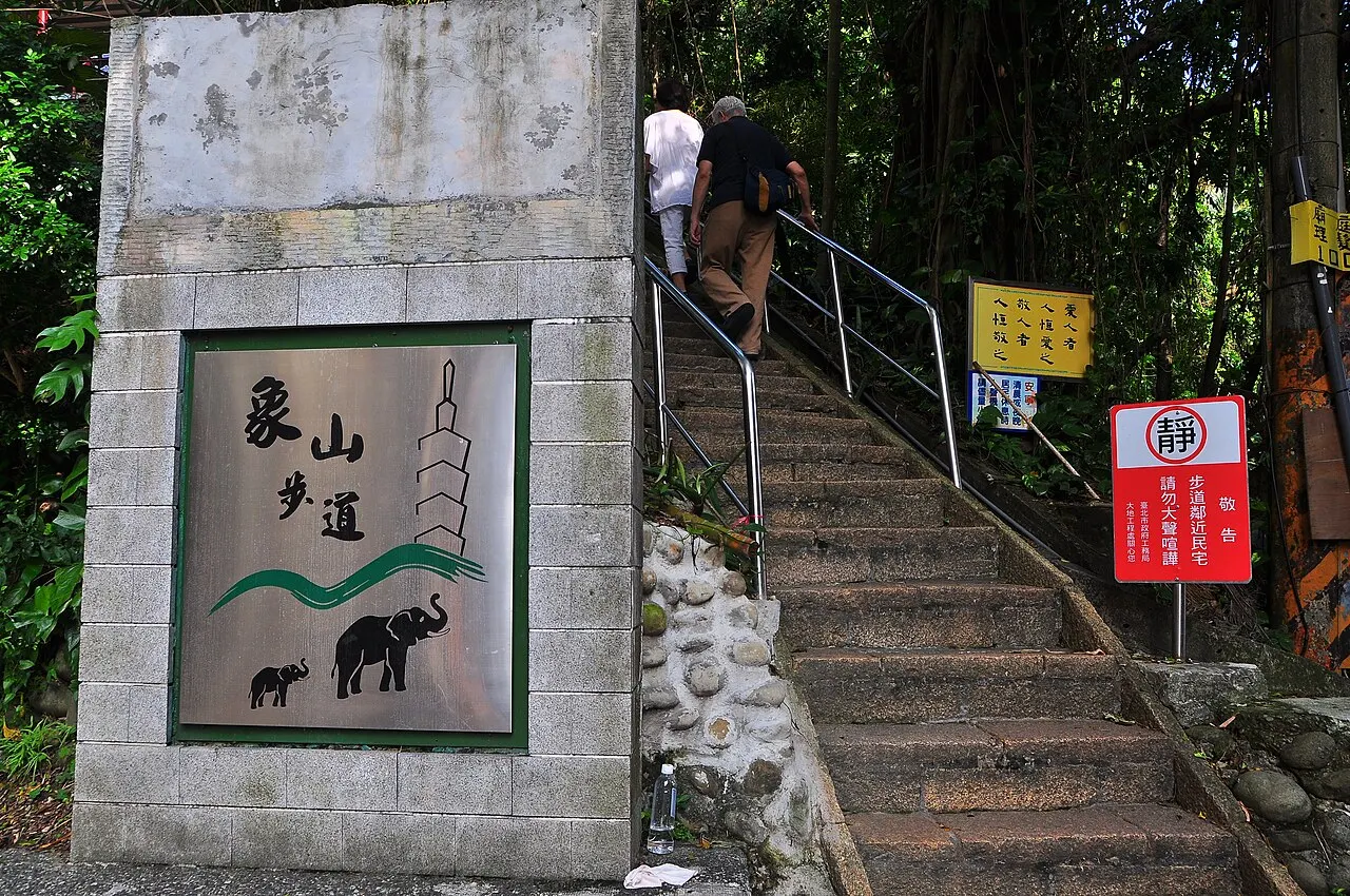 Placa retangular de pedra com o desenho de um elefante e caracteres chineses, junto a uma escadaria na mata.