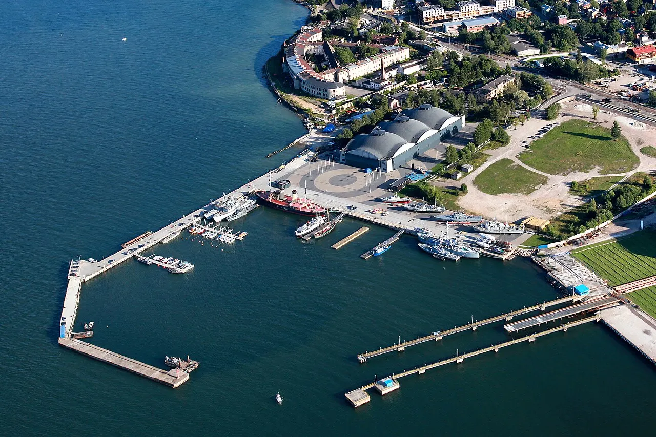 Vista aérea do Lennusadam em Tallinn: os três domos de concreto do antigo hangar de hidroaviões junto à água, com um quebra-mar em L, navios-museu atracados e o mar Báltico em volta.