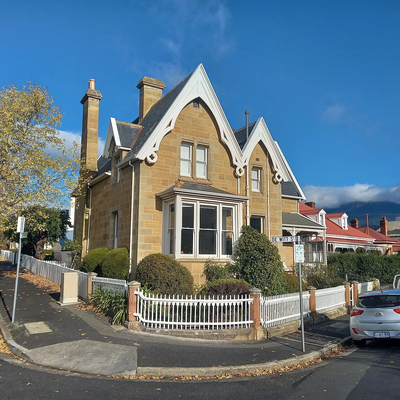 A sandstone house with white lacy timber gables and tall chimneys, behind a white picket fence on a street corner in Battery Point.