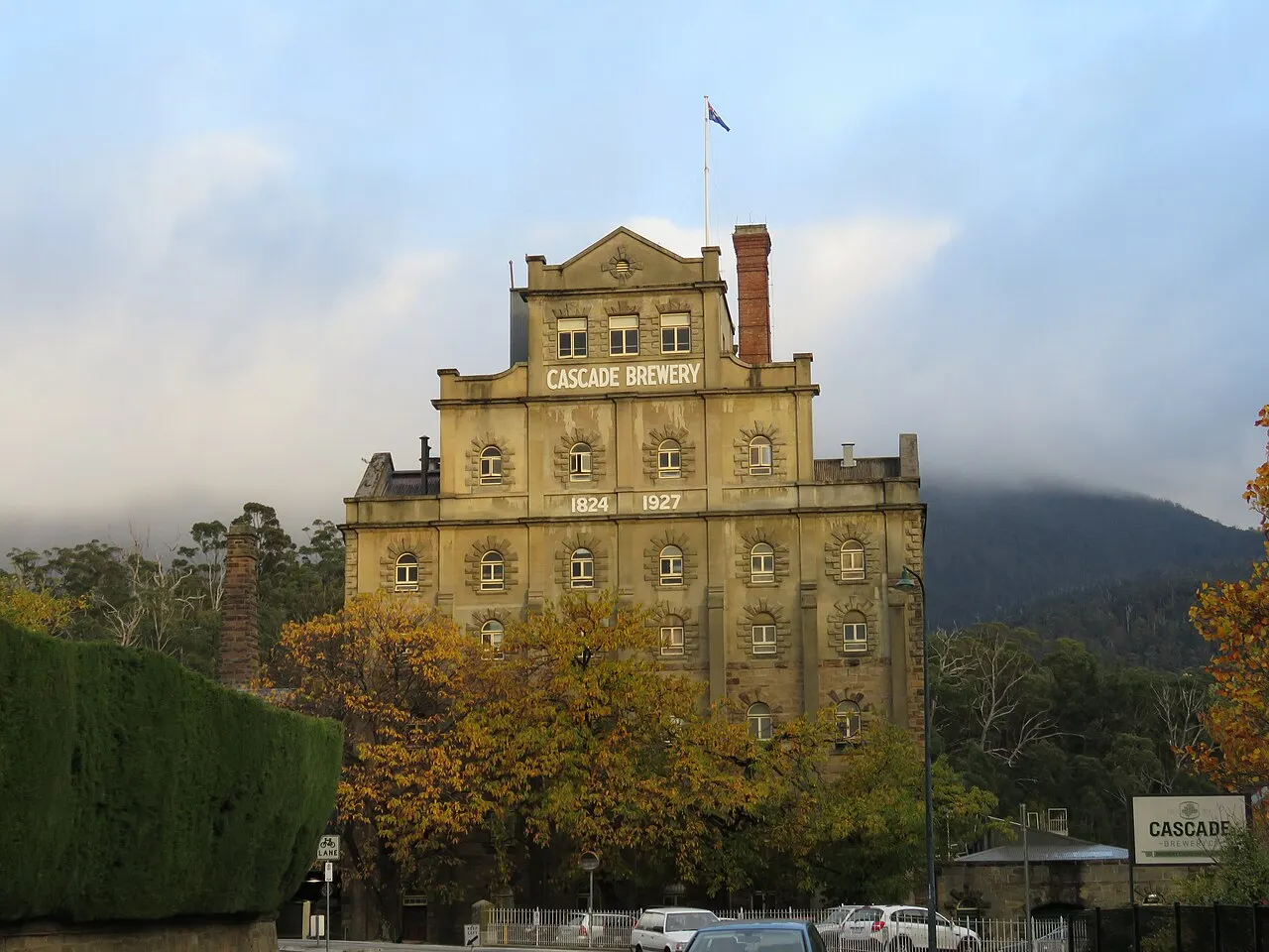 The tall sandstone Cascade Brewery building, with its name and the dates 1824 and 1927 on the facade, among autumn trees and the mist-covered hillside.