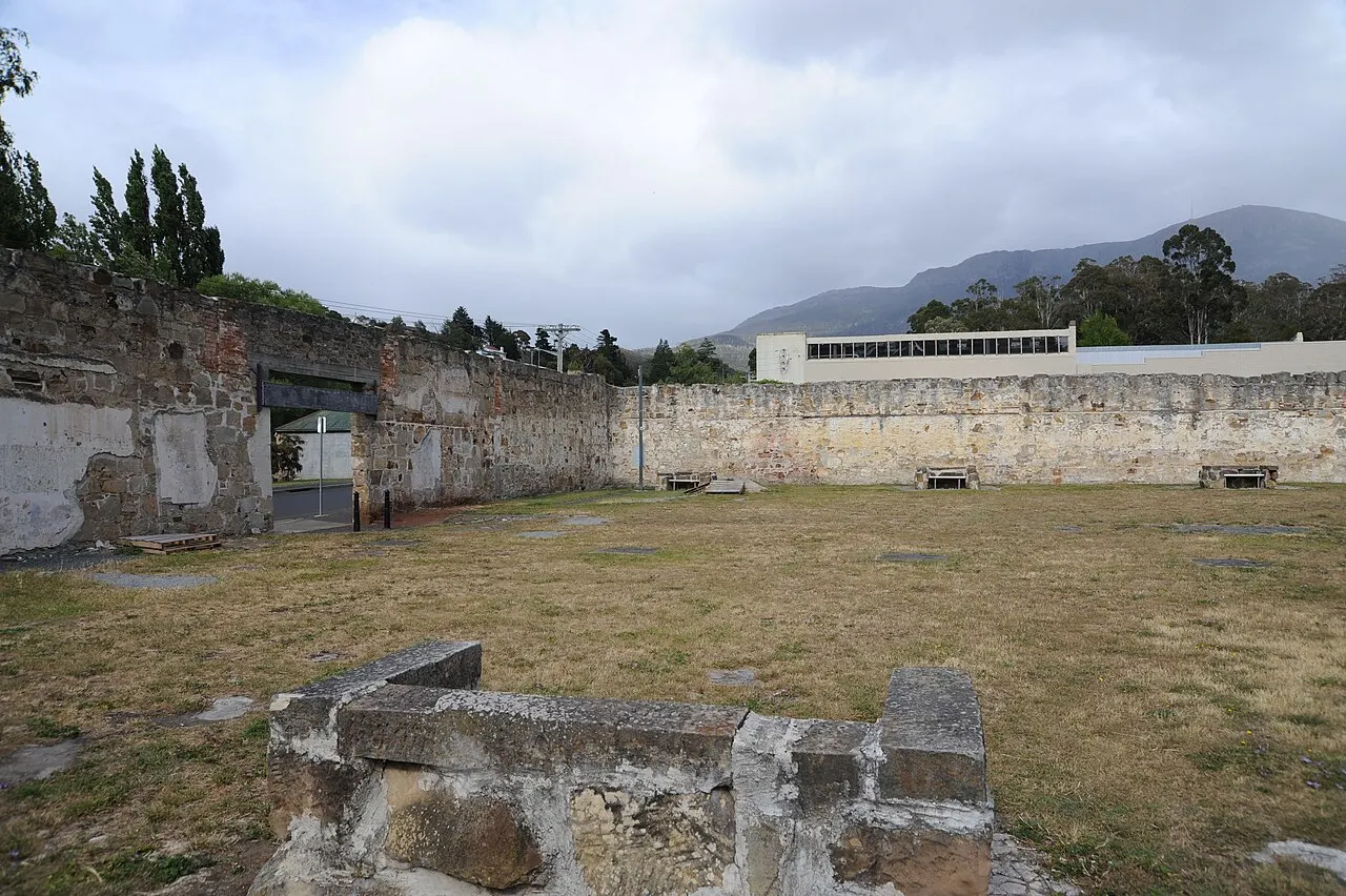 A grassy yard enclosed by high stone walls at the Cascades Female Factory, with remains of foundations in the foreground and kunanyi under cloud behind.