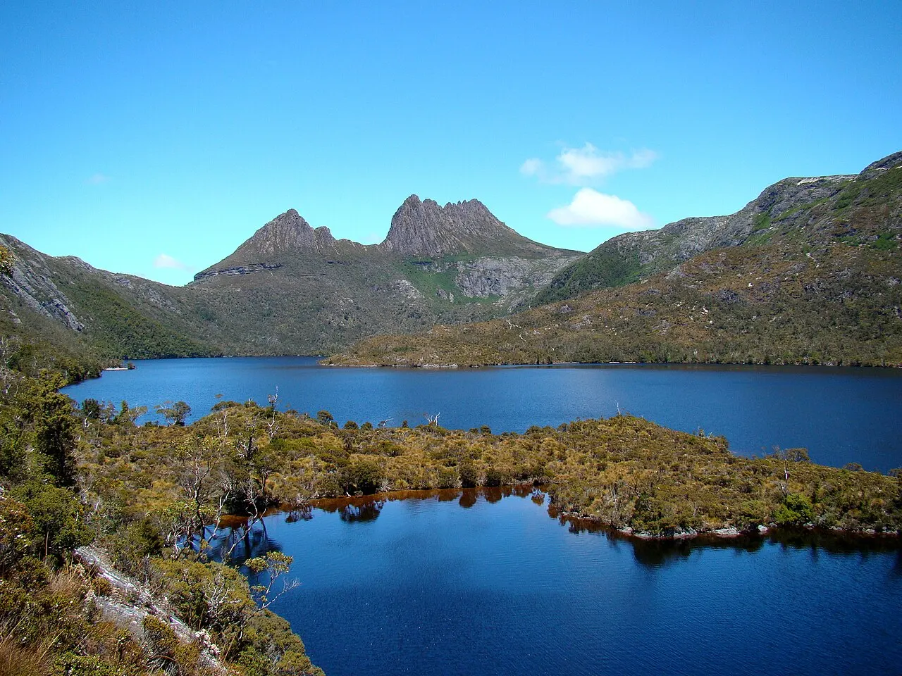The jagged peaks of Cradle Mountain behind the dark blue Dove Lake, with low alpine scrub on the shore.