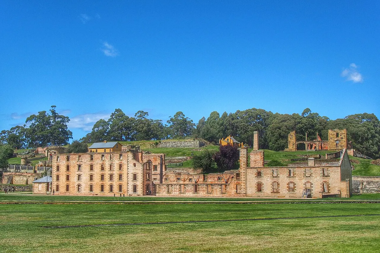 The sandstone ruins of the Port Arthur penitentiary, four storeys and roofless, on a wide lawn, with more ruins on the slope and eucalyptus trees behind.