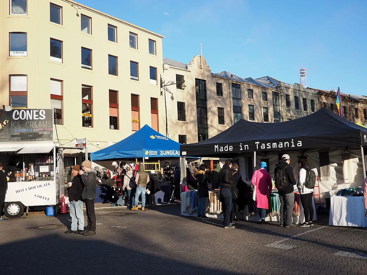 Salamanca Market stalls under blue and black awnings, an ice cream and coffee cart on the left and visitors among the tents, in front of the old buildings on the street.