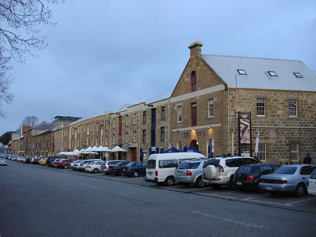 The row of sandstone warehouses on Salamanca Place at dusk, with white umbrellas on the pavement and cars parked in front.