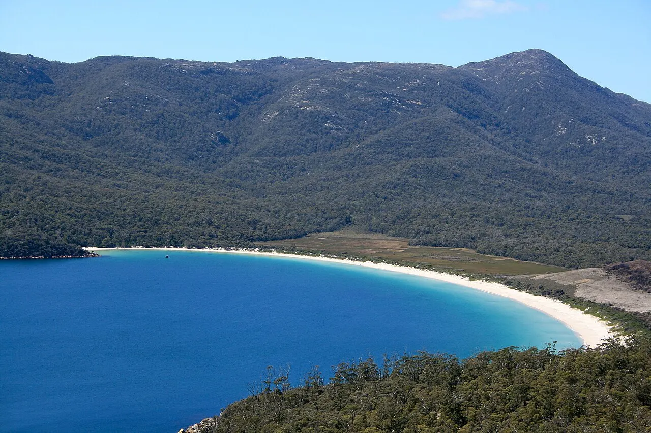 The white sand arc of Wineglass Bay between the blue water and forested mountains, seen from above.