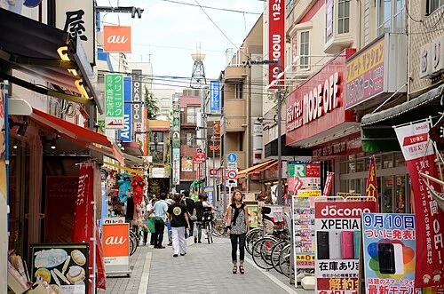 Rua estreita com letreiros, lojas e pessoas caminhando.