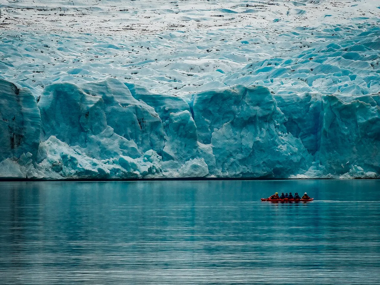 A frente de gelo azulado do Glaciar Grey sobre o lago, com um grupo de caiaques vermelhos pequeno na água diante dela.