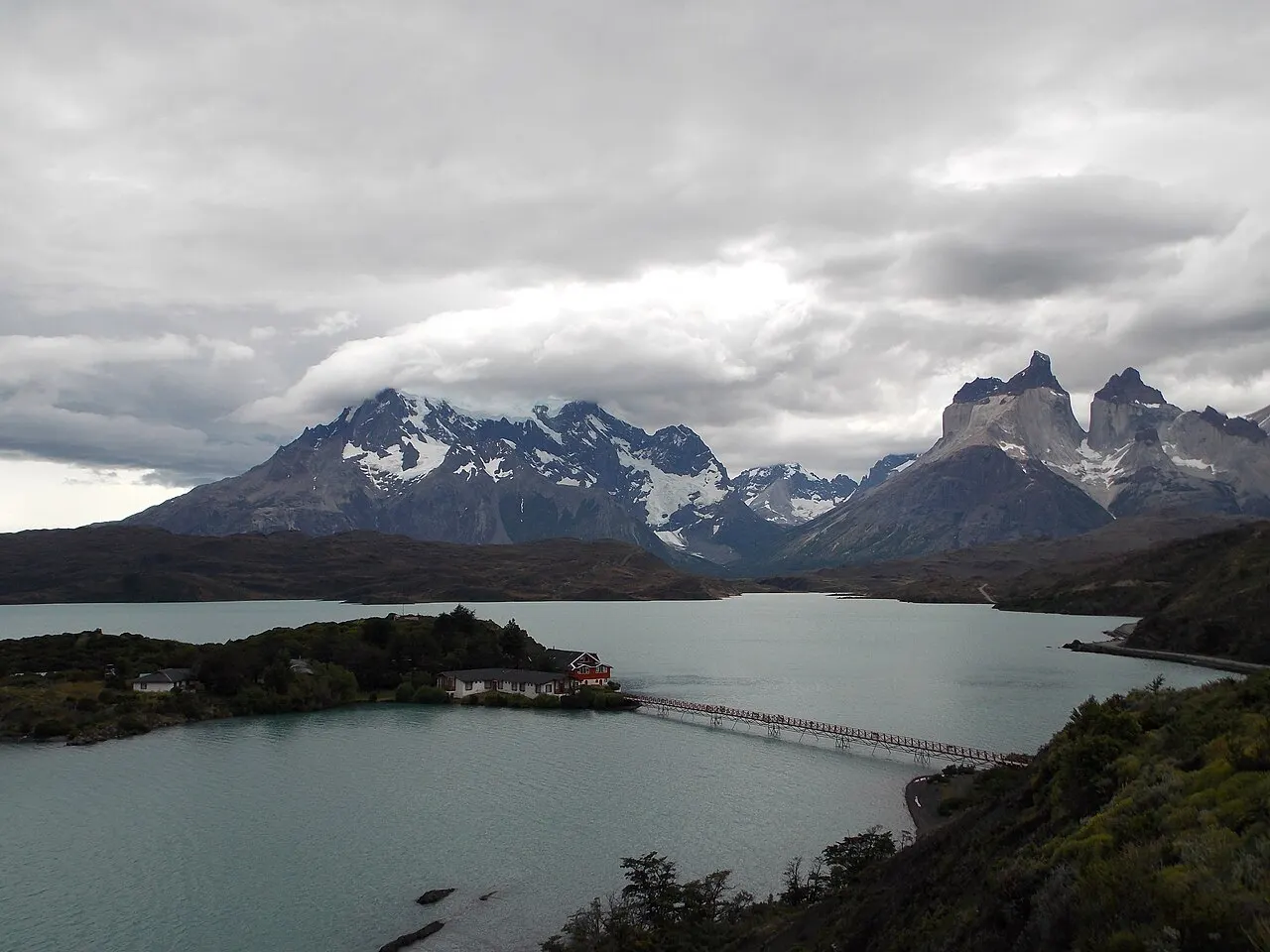 O Lago Pehoé sob céu nublado, com uma ilhota ligada à margem por uma ponte longa e os picos dos Cuernos del Paine atrás.